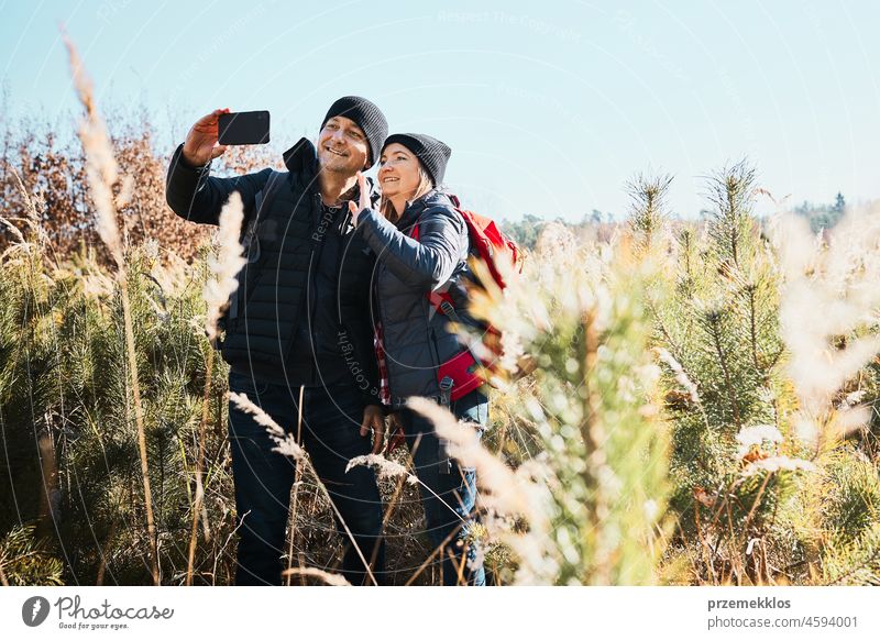 Paar, das per Videoanruf Grüße von der Urlaubsreise sendet. Wanderer mit Rucksäcken auf dem Weg in die Berge. Menschen zu Fuß durch hohes Gras entlang Weg in Wiese an sonnigen Tag