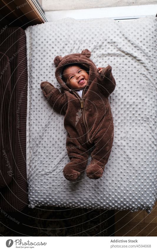 Lächelndes ethnisches Baby im weichen Bärenoverall auf einer Decke neugeboren Freude Lachen lustig spielerisch Glück expressiv gesamt bezaubernd Lügen