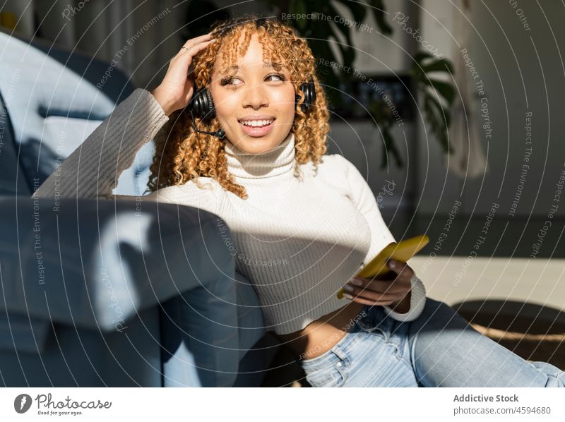 Positive Frau mit Headset und Smartphone Kopfhörer Telearbeit freiberuflich Fernarbeit Liege Stock Gerät Lächeln Musik Appartement Sonnenlicht Afroamerikaner