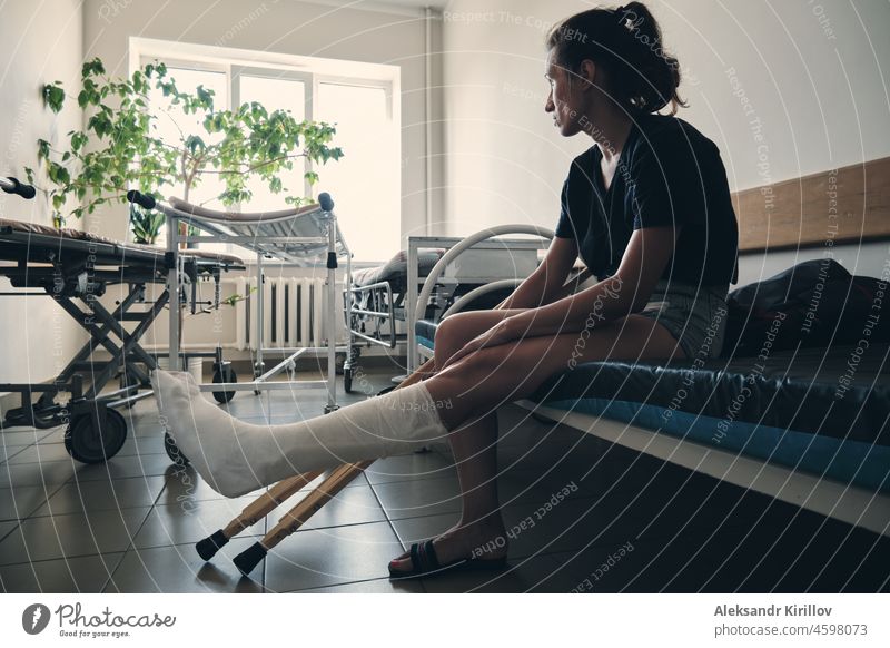 Eine Frau mit einem gebrochenen Bein sitzt auf einer Krankenhausliege und schaut aus dem Fenster. Beinverletzung. Verlust der Beweglichkeit. Rehabilitation nach einer Fraktur