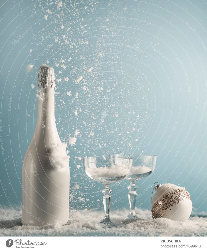 Weiße Champagnerflasche und Gläser mit Weihnachtskugel auf dem Tisch mit Schneefall auf blauen Wand Hintergrund. Neujahrskonzept mit Getränk und fallendem Schnee. Vorderansicht.