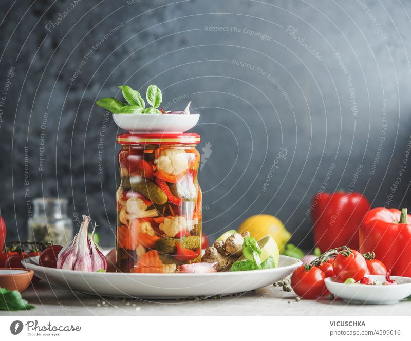 Hausgemachte Gemüsekonserven im Glas auf weißem Teller am Tisch mit Küchenutensilien und Zutaten, Kräuter und Gemüse auf schwarzem Wandhintergrund. Gesunde fermentierte Lebensmittel. Vorderansicht.