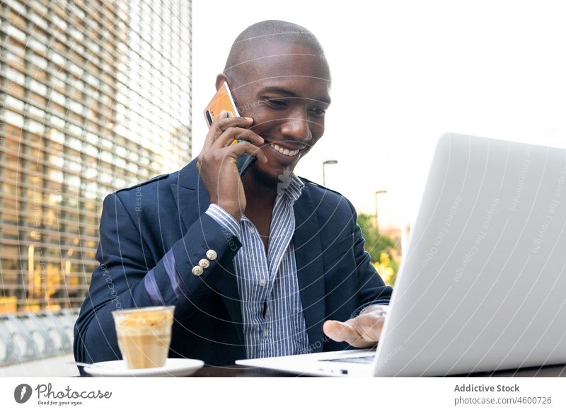 Fröhlicher schwarzer Geschäftsmann, der in der Nähe seines Laptops mit seinem Smartphone spricht Mann Unternehmer Telefonanruf Gespräch Café Terrasse Business
