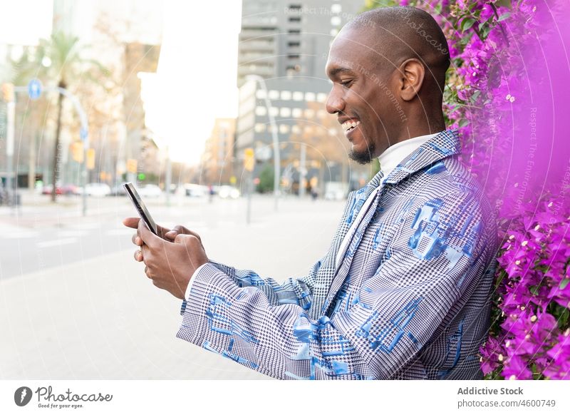 Fröhlicher schwarzer Mann, der in der Nähe von Büschen auf seinem Smartphone surft Buchse Kommunizieren Browsen Straße Pflanze Großstadt männlich Afroamerikaner