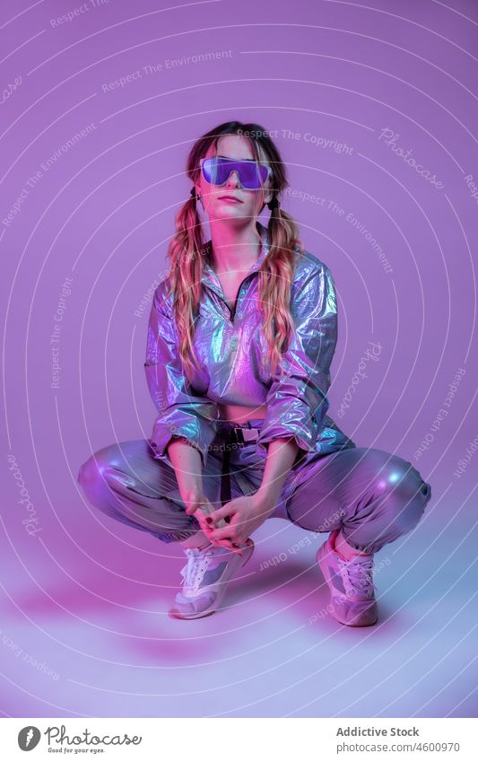 Trendige Frau mit Sonnenbrille hockt im Neonstudio neonfarbig Model leuchten Vorschein trendy Atelier Bekleidung Persönlichkeit 80s Stil futuristisch jung Stoff