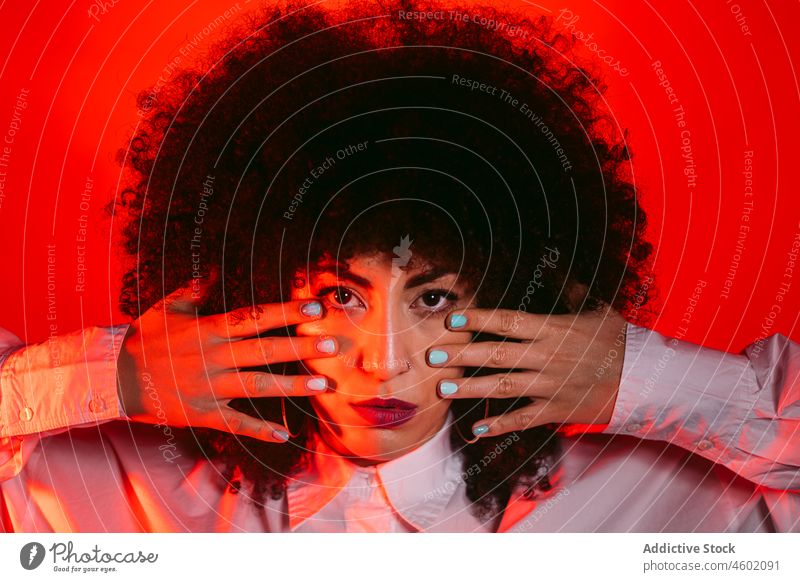 Selbstbewusste ethnische Dame, die ihr Auge mit der Hand in einem roten Studio bedeckt Frau Auge abdecken selbstsicher trendy ernst selbstbewusst Gesichtshälfte