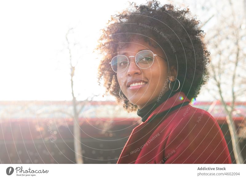 Lächelnde afroamerikanische Frau in karmesinrotem Mantel und mit Augengläsern Afro-Look Frisur positiv krause Haare Brille lässig Porträt heiter Sonnenlicht