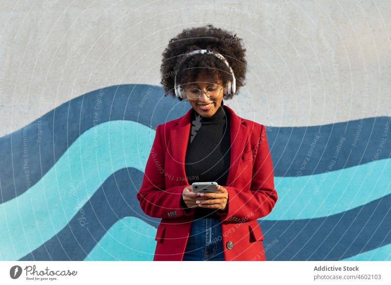 Stilvolle schwarze Frau mit Brille, die mit Kopfhörern Musik hört Afro-Look Smartphone zuhören Rollkragenpulli Browsen Drahtlos Podcast benutzend Talkrunde