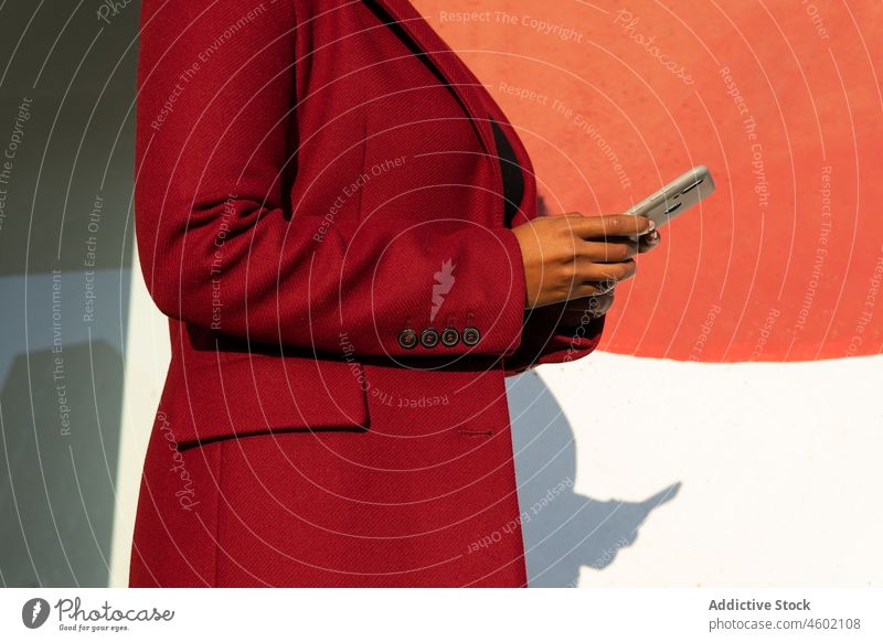 Anonyme afroamerikanische Frau in formeller Jacke mit Smartphone Apparatur Funktelefon benutzend Gerät Straße Browsen rot Sonnenlicht Talkrunde lässig Surfen