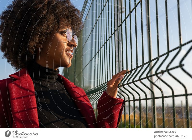 Schwarze Frau mit Afrofrisur schaut durch Metallgitter weg Afro-Look Zaun Raster Frisur Straße formal selbstbewusst Stil Vorschein Outfit krause Haare trendy
