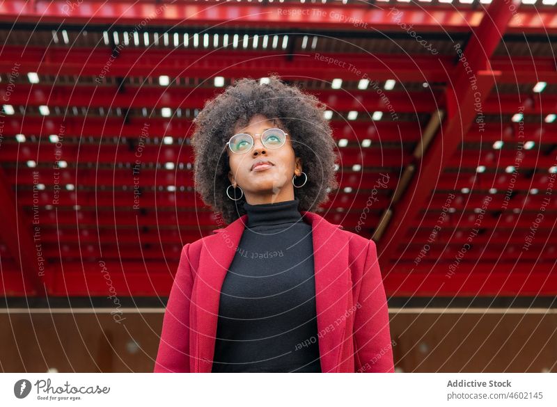 Afroamerikanische Frau in karmesinrotem Mantel und mit Brille Afro-Look Frisur positiv krause Haare lässig Porträt charmant Optimist Menschliches Gesicht