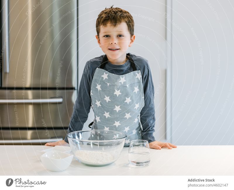 Lächelnder Junge mit einer Schüssel Mehl in der Küche Koch kulinarisch Kind Bestandteil heimwärts heimisch vorbereiten Schalen & Schüsseln Tisch Schürze heiter
