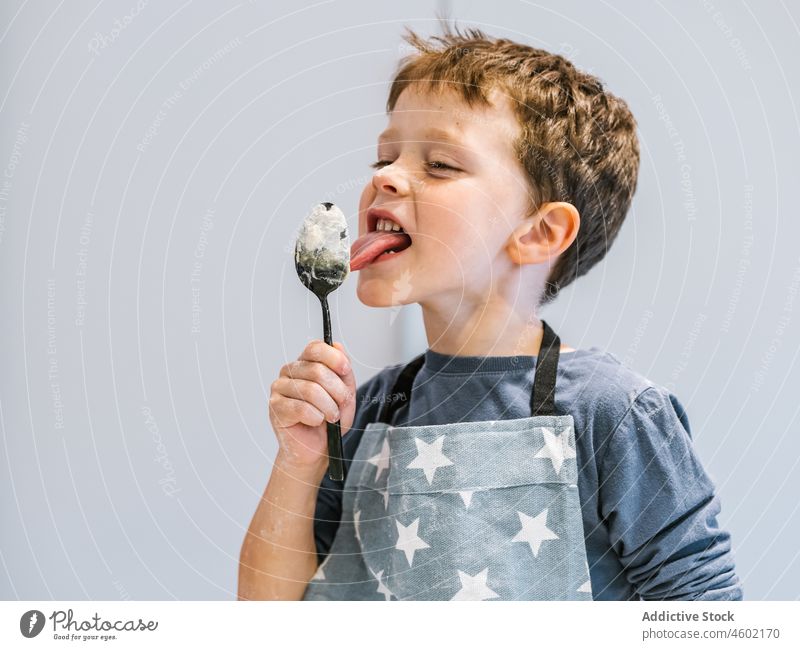Junge in Schürze leckt Löffel mit Teig Koch Kind kulinarisch lutschen heimisch Küchengeräte Helfer zu Hause dreckig Utensil lustig vorbereiten unordentlich