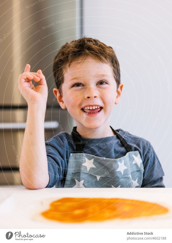 Lächelnder Junge in Schürze in der Küche Koch heimwärts Kind heimisch vorbereiten Küchengeräte kulinarisch braune Haare blaue Augen zu Hause charmant niedlich