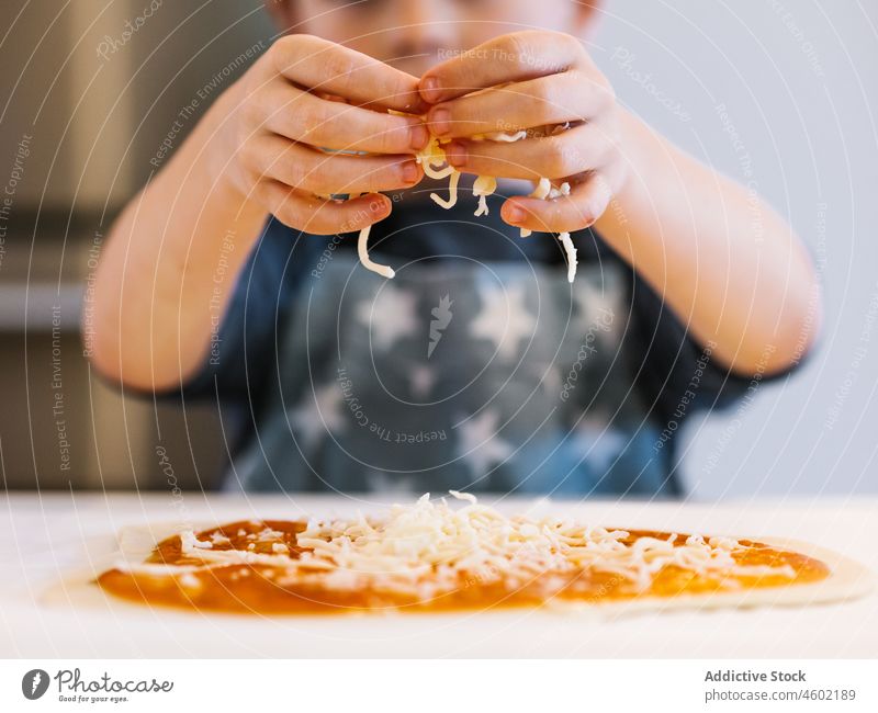 Anonymer Junge belegt Pizza mit Käse Koch Kind kulinarisch Küche Teigwaren Helfer Küchengeräte heimisch zu Hause Schürze Rezept Lebensmittel hinzufügen