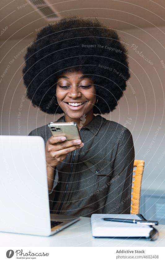 Fröhliche schwarze Frau, die in der Nähe ihres Laptops auf ihrem Smartphone surft Arbeit Textnachricht Browsen online freiberuflich Arbeitsplatz Internet