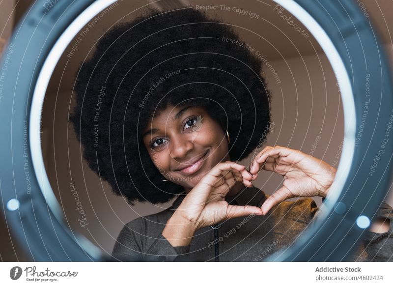 Lächelnde schwarze Frau zeigt Herzgesten durch Ringlampe Blogger gestikulieren Liebe Vlogger Freizeit Afro-Look Frisur feminin Vorschein Glück charmant brünett