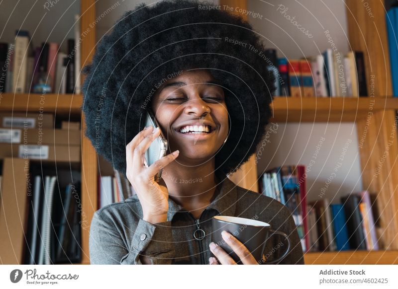 Fröhliche schwarze Frau mit Kaffee, die mit ihrem Smartphone spricht Telefonanruf Gespräch diskutieren Heißgetränk Getränk Afro-Look Lächeln Talkrunde Freizeit