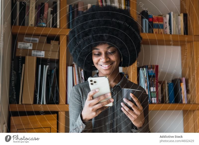 Fröhliche schwarze Frau mit Kaffee, die mit ihrem Smartphone spricht Gerät Textnachricht Heißgetränk Getränk Afro-Look Lächeln Talkrunde Freizeit Browsen online
