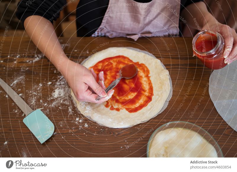Anonyme Frau gibt Soße auf den Teig Koch Teigwaren Pizza Saucen Tomate Küche kulinarisch selbstgemacht roh vorbereiten Küchenchef Tisch Schürze Prozess Licht