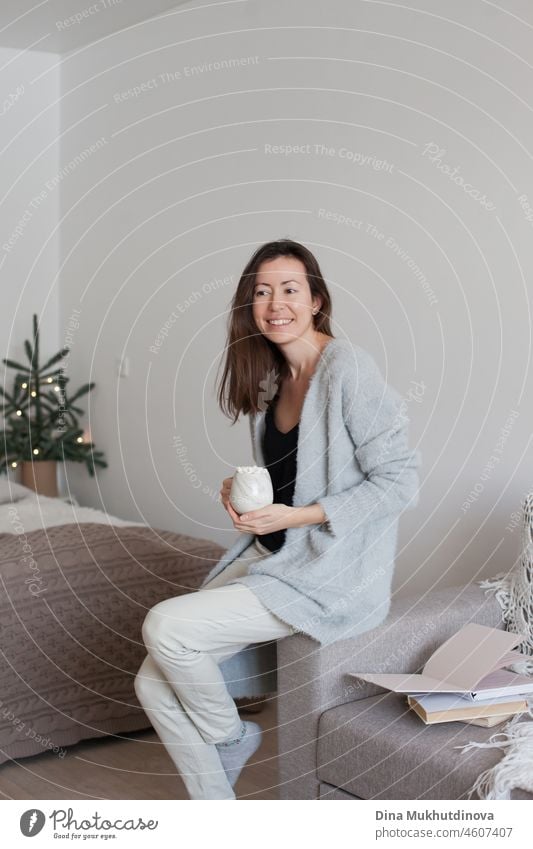 Millennial Frau in gemütlichen Wohnung hält eine Tasse Kaffee sitzen auf der Couch in der Nähe von offenen Bücher auf Urlaub Saison Lifestyle tausendjährig