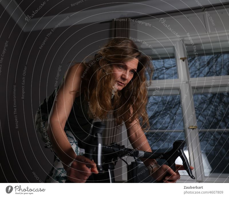 Sportliche schöne Frau trainiert auf Indoor-Rennrad, dahinter Fenster mit Abendlicht sportlich trainieren Gute Vorsätze Heimtrainer Rennlenker grün neon Fitness
