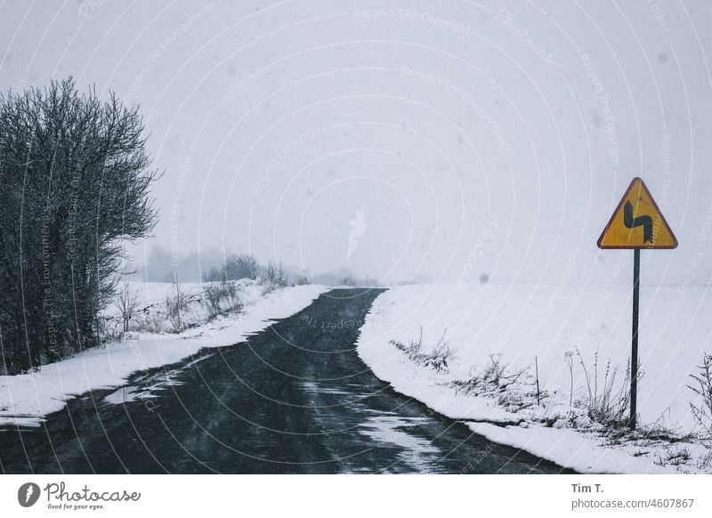 ein Schild warnt an einer winterlichen kleinen Strasse vor einer Kurve. Landstraße Winter polska Polen Schnee Asphalt Verkehrszeichen Verkehrsschild Straße