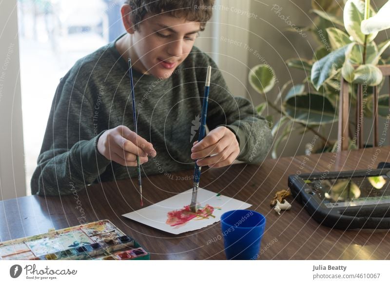 Junge mit besonderen Bedürfnissen malt mit beiden Händen gleichzeitig Aquarelle; in der Nähe sitzt ein Kommunikationsgerät für die Unterstützte Kommunikation