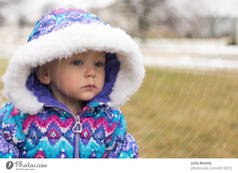 Ein 18 Monate altes Mädchen in einem bunt gemusterten Schneeanzug mit Kapuze schaut mit ernster Miene von der Kamera weg und wartet darauf, dass der Schnee fällt.