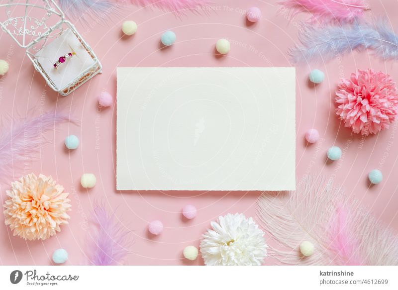 Blanko-Karte zwischen pastellfarbenen Blumen, Bommeln und Federn in der Nähe von Ring in einer Geschenkbox auf rosa Hochzeit Kuvert Pompom Attrappe Draufsicht