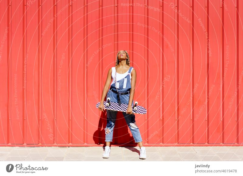 Schwarzes Mädchen gekleidet lässig, wtih ein Skateboard auf roten städtischen Wand Hintergrund. schwarz Zopf afrobraid Rastalocken Frau Afrikanisch Frisur