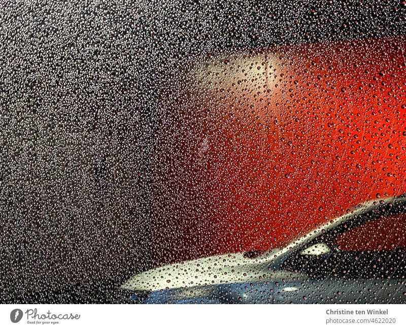 Blick durch ein verregnetes Autofenster, nebenan parkt ein silbernes Auto vor einer roten beleuchteten Wand Regenwetter Regentropfen Fenster rote Wand parken