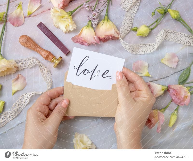 Hände mit Karte LOVE im Umschlag in der Nähe von rosa Blumen auf einem Marmortisch Postkarte handschriftlich mich heiraten immer Vorschlag Heirat Draufsicht