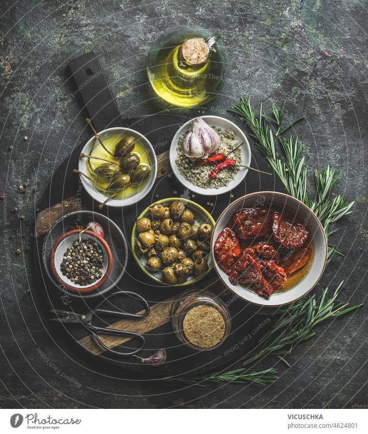 Verschiedene leckere mediterrane Vorspeisen in Schalen: Oliven, Kapern, getrocknete Tomaten, frische Rosmarinkräuter und Olivenöl, serviert auf einem rustikalen Schneidebrett