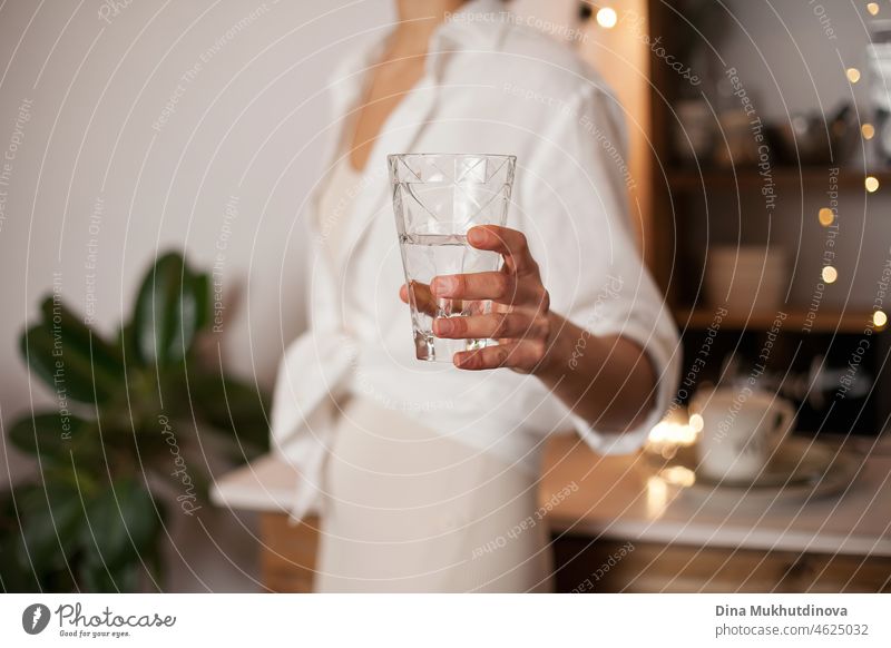 Unbekannte Frau in weißem Hemd mit einem Glas Wasser in der Hand, stehend in der Küche bei Abendlicht. Gesunder Lebensstil - jeden Tag ausreichend Wasser trinken