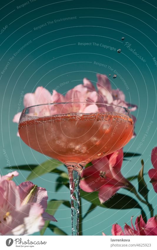 Nahaufnahme von Wein oder Cocktail in einem Glas, umgeben von rosa und magentafarbenen Alstroemeria-Blüten auf blauem Grund trinken Getränk alkoholisch kalt