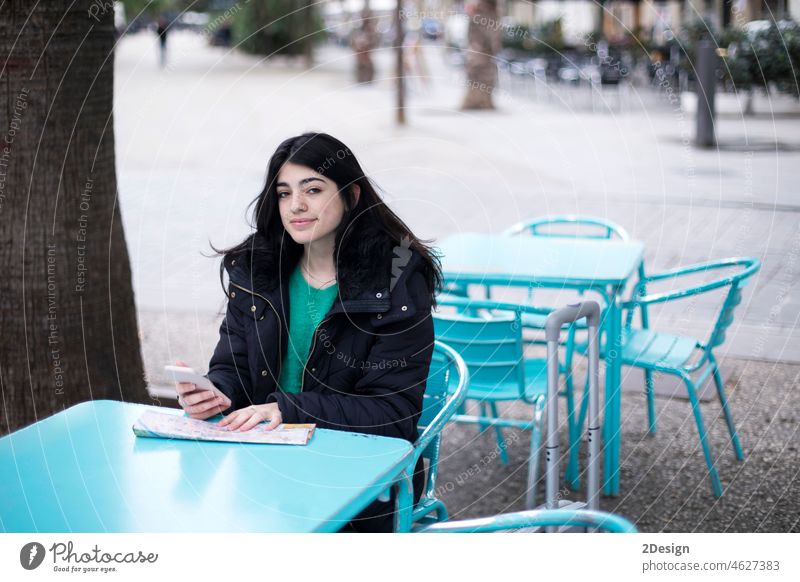 Junge Touristin sitzt in einem Straßencafé am Tisch und sucht auf einer Karte nach einem Weg, wobei sie ihr Smartphone benutzt. Frau Mobile Café Sitzen