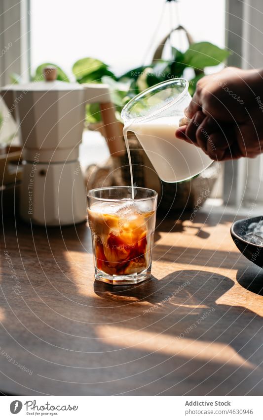 Anonyme Person, die Milch in ein Glas mit Kaffee und Eis gießt melken kalt Eiswürfel Kaffeepresse eingießen Koffein Getränk Erfrischung Küche Morgen Tisch