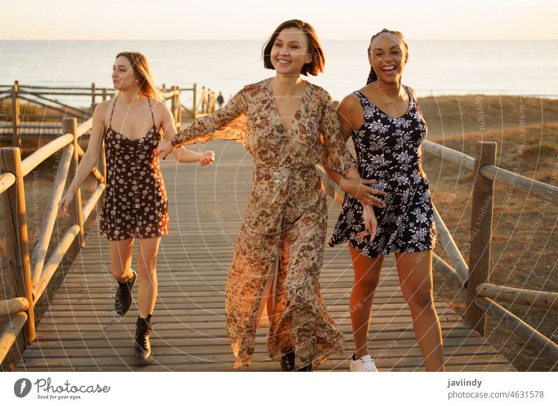 Lächelnde multiethnische Freundinnen, die auf der Holzpromenade am Meer spazieren gehen Frauen Freude heiter Sonnenuntergang Promenade Stil Küste Glück