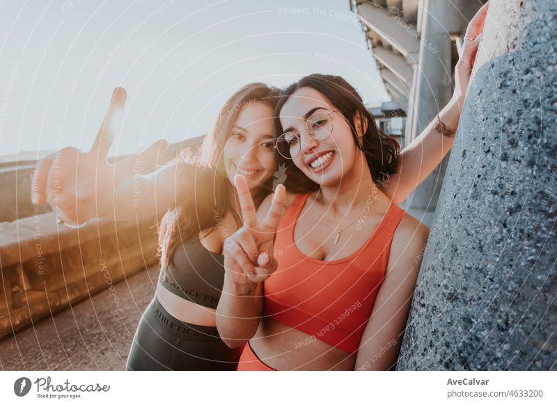 Zwei Junge fröhliche Sportlerin Training im Freien tun Übung lächelnd mit glücklich in die Kamera tun Sieg Zeichen mit den Fingern während einer bunten Sonnenuntergang. Working out zusammen Konzept. Gesundes Leben