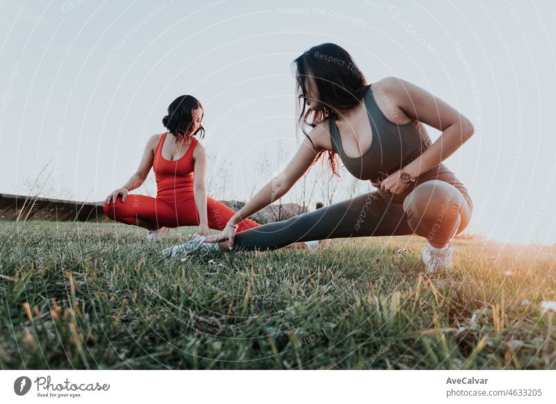 Zwei junge afrikanische Frauen strecken ihre Beine auf dem Gras während eines Sonnenuntergangs, sportliche Frau vor dem Laufen. Training im Park nach der Arbeit, Training in der Stadt, Spaß zusammen haben