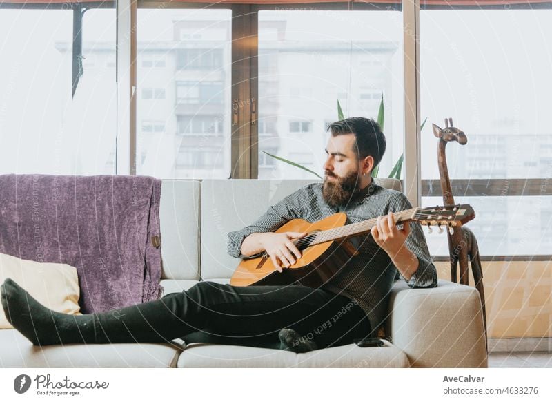 Junger hübscher bärtiger Mann spielt akustische spanische Gitarre auf dem Sofa im Wohnzimmer. Lernen, ein neues Instrument zu spielen. Neue Gewohnheit Konzept, Fähigkeiten und Hobby. Modernes Zuhause trendige Kleidung