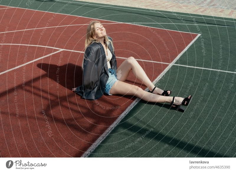 Junge blonde Frau in Stöckelschuhen und Jacke, sitzend auf einem Backetball-Sportplatz alleinstehende Erwachsene attraktiv schön Lächeln Glück Windstille