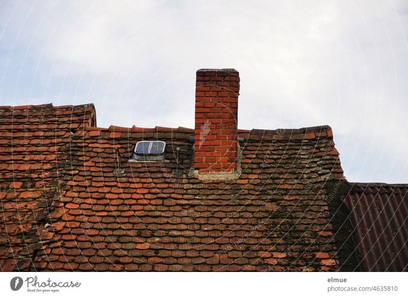 altes Ziegeldach eines Reihenhauses mit einem unverputzten Schornstein, einem kleinen Dachfenster und angrenzendem Wellblechdach / wohnen rot Biberschwanzziegel