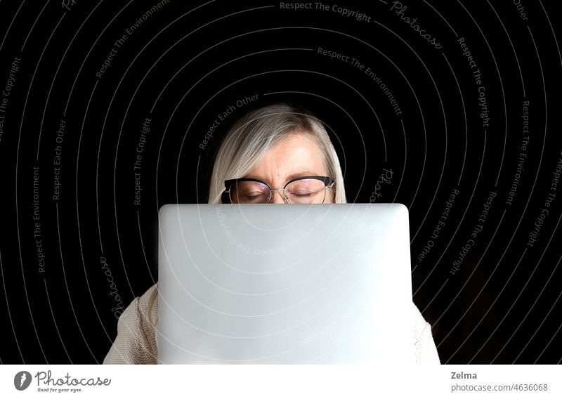 müdes reifes Frauengesicht mit geschlossenen Augen und Brille hinter einem Computer-Laptop versteckt, isoliert auf einem schwarzen Hintergrund Gesicht heimwärts