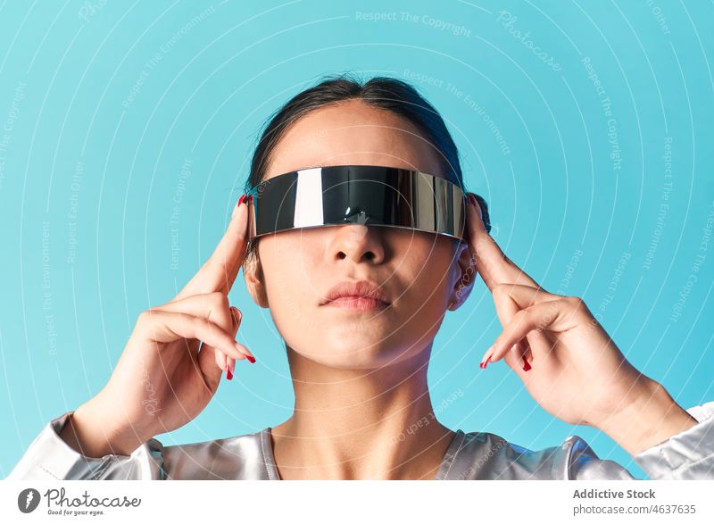 Selbstbewusste junge ethnische Frau mit verspiegelter VR-Brille im blauen Studio Virtuelle Realität Erfahrung Stil futuristisch erweitert Porträt unterhalten