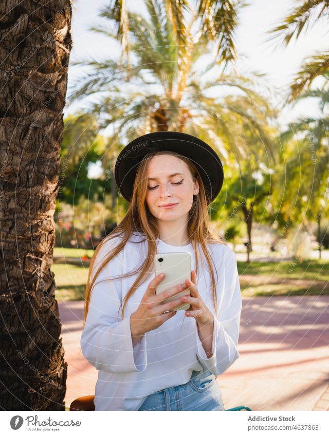 Junger Radfahrer benutzt Smartphone im Park Frau benutzend soziale Netzwerke Sommer Handfläche fettarm jung Baum Blume ruhen Stil Hut tagsüber lässig Browsen