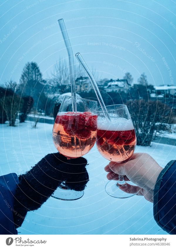 Zwei Hände, die mit einem kalten. alkoholischen Getränk mit Himbeeren und Glas-Strohhalm im Schnee anstoßen - Bromskirchen, Nordrhein-Westfalen, Deutschland