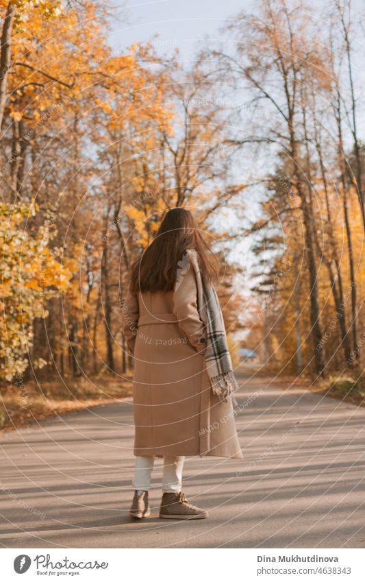 Junge brünette Frau mit langen Haaren in beige Mantel zu Fuß im Herbst Park auf einer Straße. Entspannende Momente in der Natur, Millennial Mädchen glücklich zu sein und lächelnd. Frau von hinten zu Fuß im Herbst Park.