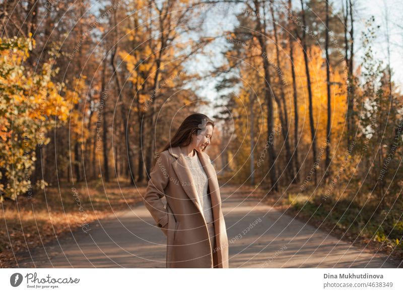 Junge brünette Frau mit langen Haaren in beige Mantel zu Fuß im Herbst Park auf einer Straße. Entspannende Momente in der Natur, Millennial Mädchen glücklich zu sein und lächelnd. Frau lächelnd zu Fuß im Herbst Park.